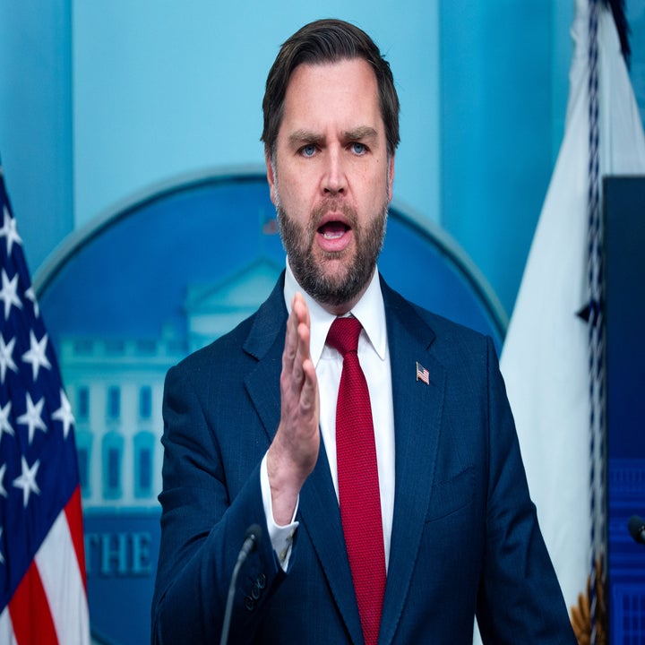 Person i dress og rødt slips snakker på en pressekonferanse, gestikulerer med én hånd, mot et bakteppe som ligner en regjeringssetting