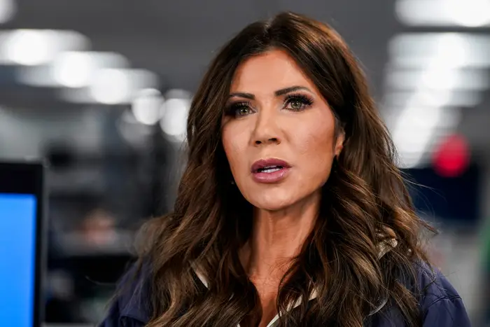 En person snakker på et innendørs arrangement, iført en mørk jakke med stylet hår