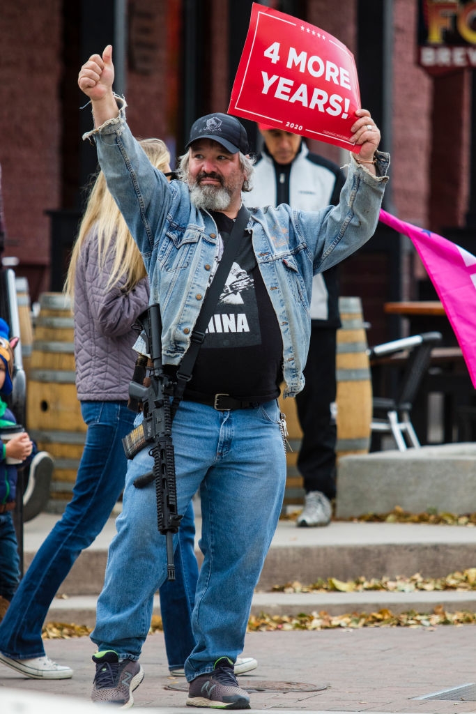 Mann i dongerijakke og caps holder "4 More Years!" tegn, gir tommelen opp. Han har et skytevåpen slengt over brystet