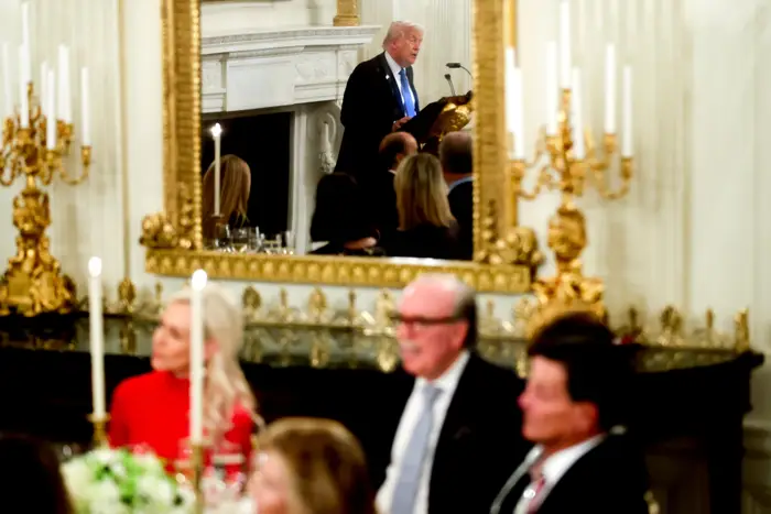 Mann snakker ved et podium i en formell arrangementssal med et utsmykket speil. Gjestene sitter ved bordene i forgrunnen