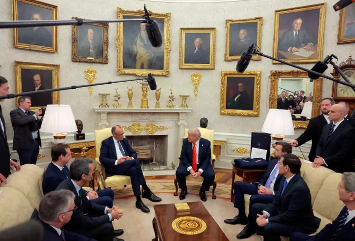 Et møte i en formell kontorsetting med folk sittende og omgitt av portrettmalerier og mikrofoner over hodet