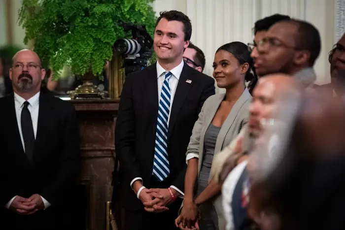 En gruppe mennesker på et arrangement, med noen i formell antrekk. En kvinne i grå blazer og svart topp står ved siden av en mann i dress og stripet slips