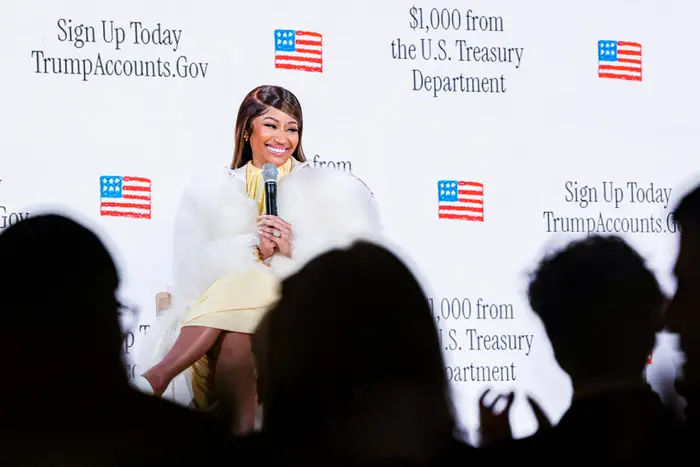 En person sitter på scenen iført en elegant hvit pels over en elegant kjole, og snakker på et salgsfremmende arrangement med et bakteppe som viser tekst om det amerikanske finansdepartementet