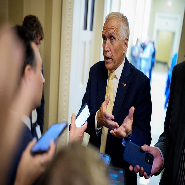 En mann i dress snakker med flere journalister som holder telefoner og mikrofoner i en gang