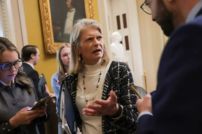 Kvinne i mønstret blazer og perlekjede snakker med journalister som holder notatbøker og telefoner i en innendørs setting med kunstverk i bakgrunnen