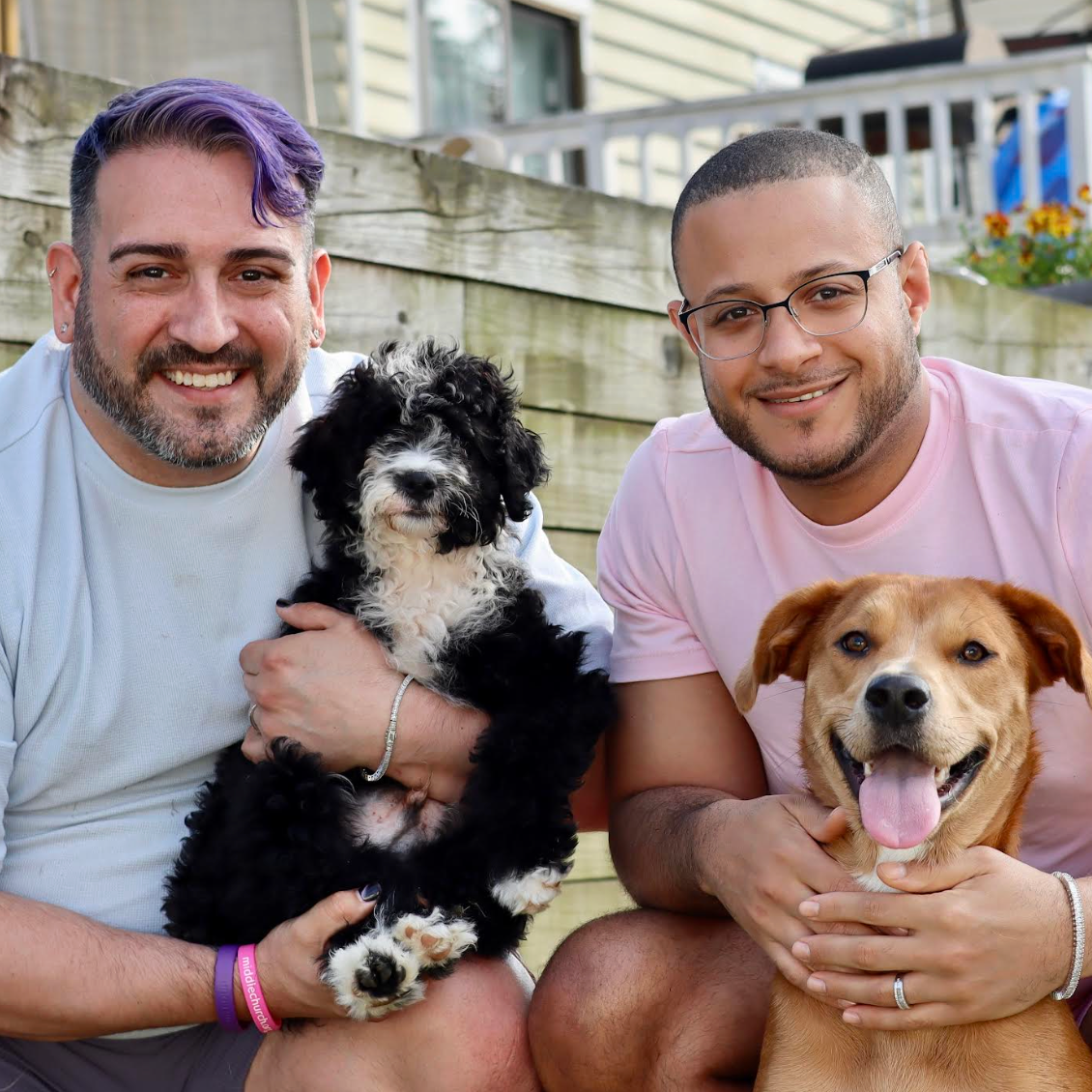 To personer som smiler mens de holder to hunder i utendørs omgivelser. De kneler på gresset med et tregjerde og hus i bakgrunnen