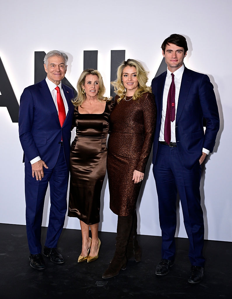 Fire personer poserer sammen på et formelt arrangement. Tre bruker dresser, en har på seg en skinnende kjole med støvler. De står foran en vegg med stor tekst