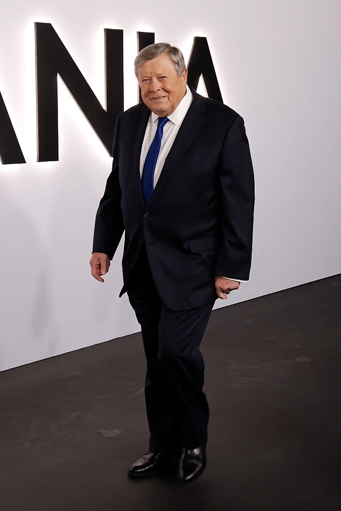 Mann i formell dress med blått slips stående foran en vegg med delvis tekst synlig