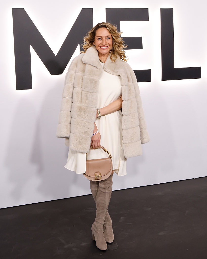 Person i hvit kjole og pelsjakke med knehøye semskede støvletter poserer smilende på en rød løper foran et stort skilt