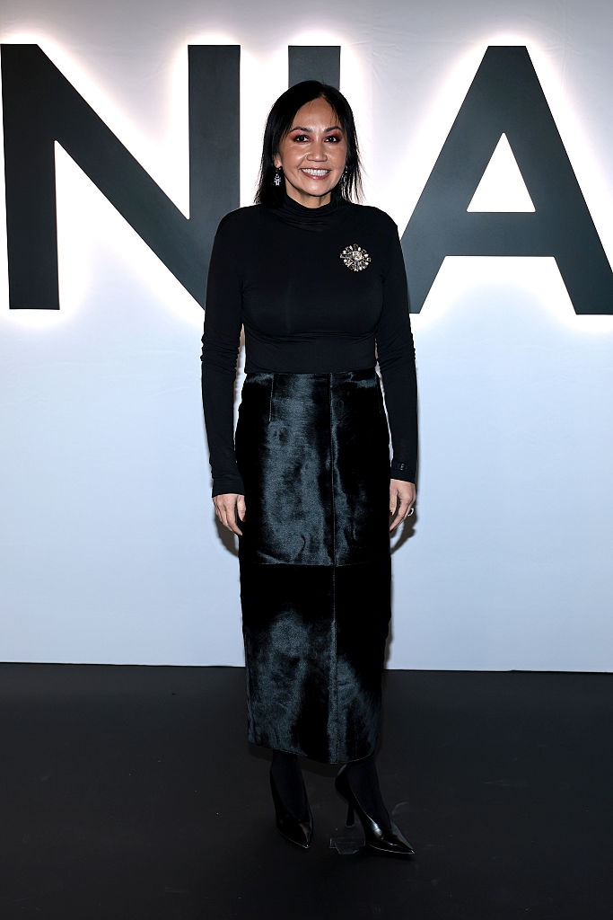 Person i elegant antrekk med langermet topp og knelangt skjørt, stående foran et stort skilt på et kjendisarrangementsteppe