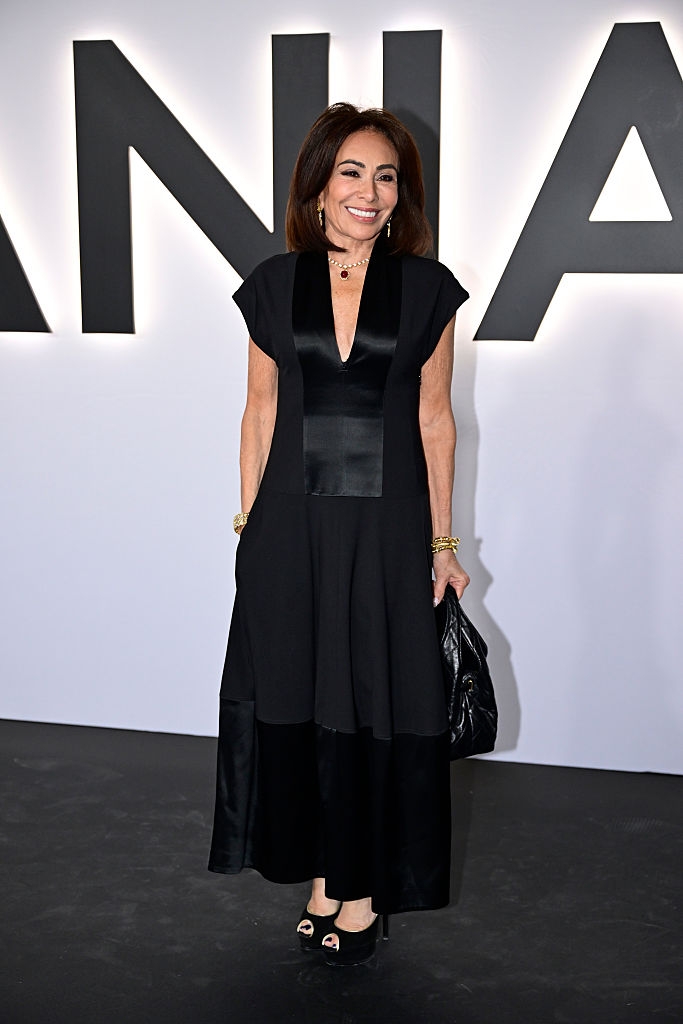 En kvinne i en elegant svart kjole med dyp V-hals står på en rød løper, poserer med et smil og holder en clutchveske