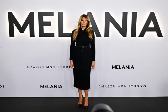 Person i elegant, langermet svart kjole poserer på et arrangement med "Melania" og "Amazon MGM Studios" synlig på bakgrunnen