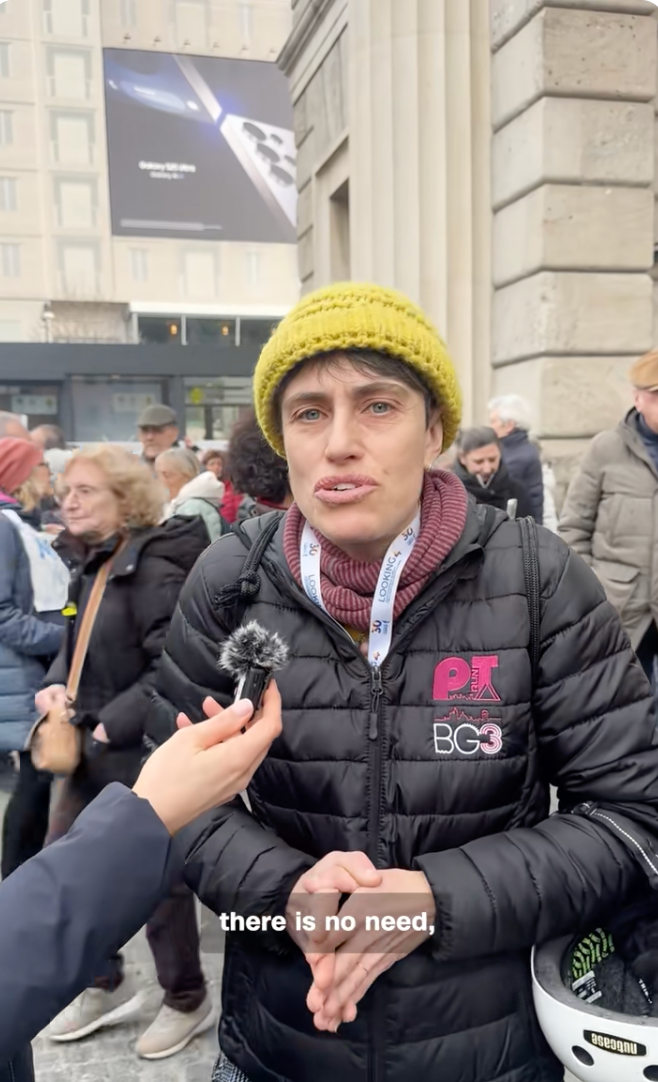 En person som har på seg en strikkelue og pufferjakke snakker under et gateintervju. Folk er samlet i bakgrunnen. Undertekster lest, 