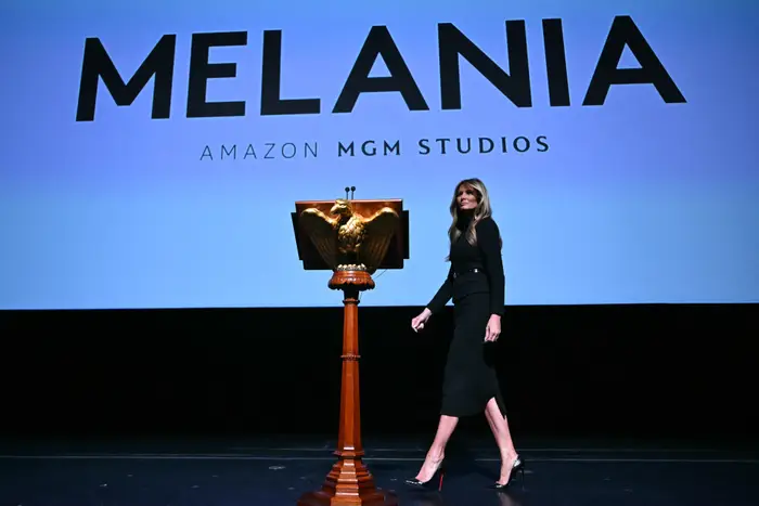 Person i en elegant svart kjole står på scenen ved siden av et podium med et ørneemblem. "MELANIA" og "Amazon MGM Studios" vises i bakgrunnen