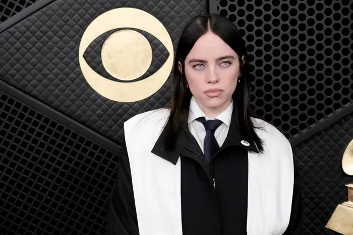 Person i et formelt ensemble med slips og svart-hvitt jakke står på en mediebegivenhet i bakgrunnen med en fremtredende logo