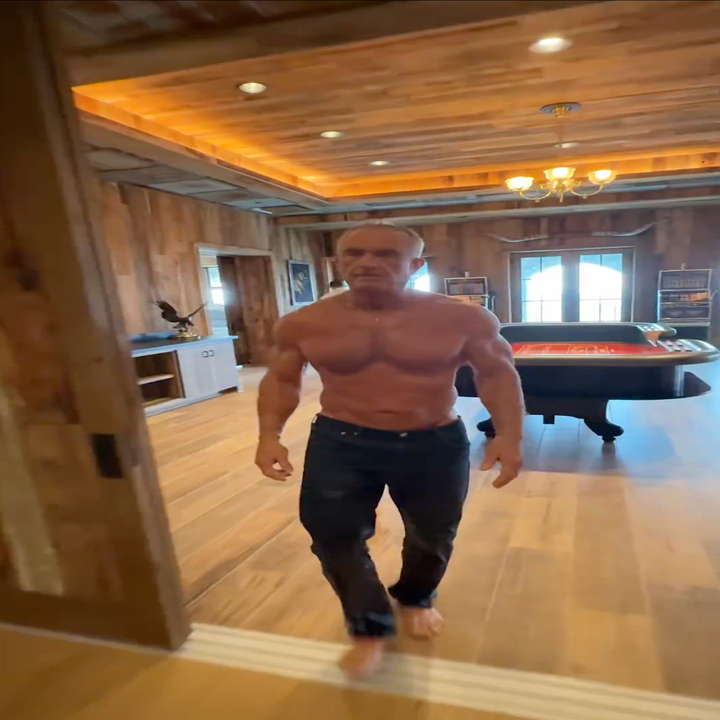 Mann uten skjorte med muskuløs kropp går barbeint i et rom med trepanel og et biljardbord i bakgrunnen