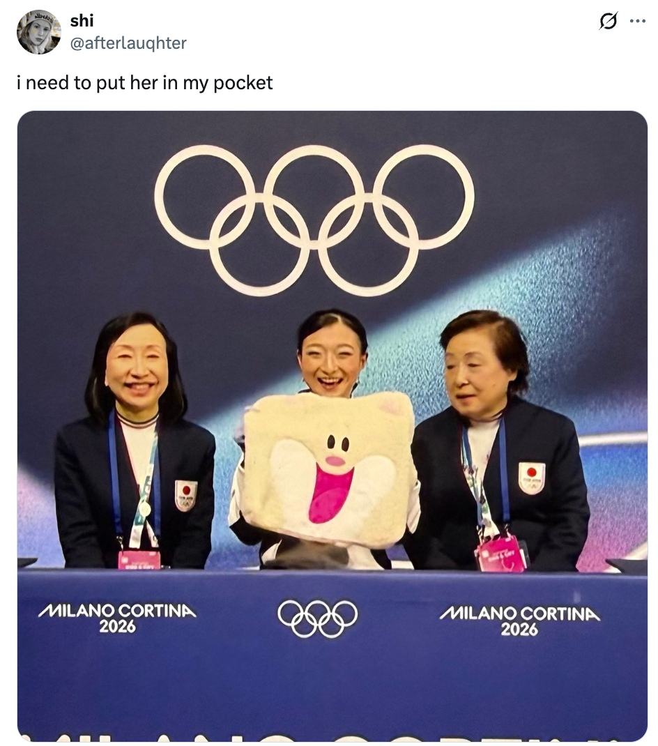 Tre personer sitter ved et bord foran Milano Cortina 2026 OL-logoer og holder en plysj med et glad ansikt