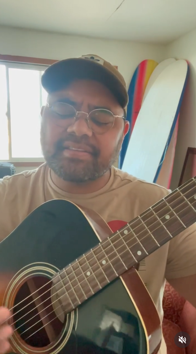 Person i uformelle omgivelser som spiller en akustisk gitar, iført hette og briller, med surfebrett synlige i bakgrunnen