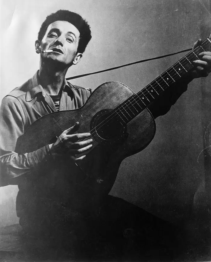 Person som spiller en akustisk gitar med en sigarett i munnen, iført en uformell skjorte, ser oppover