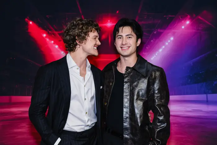 To personer som smiler til hverandre mens de står på en arena. Den ene har på seg dress, den andre en skinnende jakke. Atmosfæren er levende og livlig