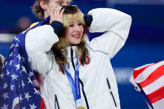 Idrettsutøver på podiet iført en hvit jakke holder et amerikansk flagg og gullmedalje, smilende under OL-seierseremoni