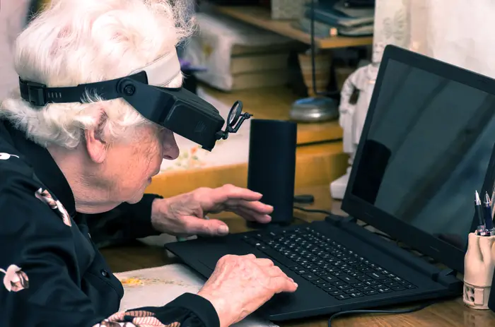 Eldre voksen bruker en skjermforstørrelsesglass for å se en bærbar datamaskin, sittende ved et skrivebord med kontorrekvisita