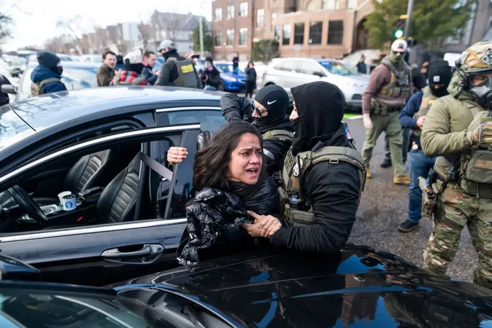 En kvinne blir tvunget fjernet fra en bil av enkeltpersoner i taktisk utstyr, midt i et kaotisk gatebilde med tilskuere og polititilstedeværelse