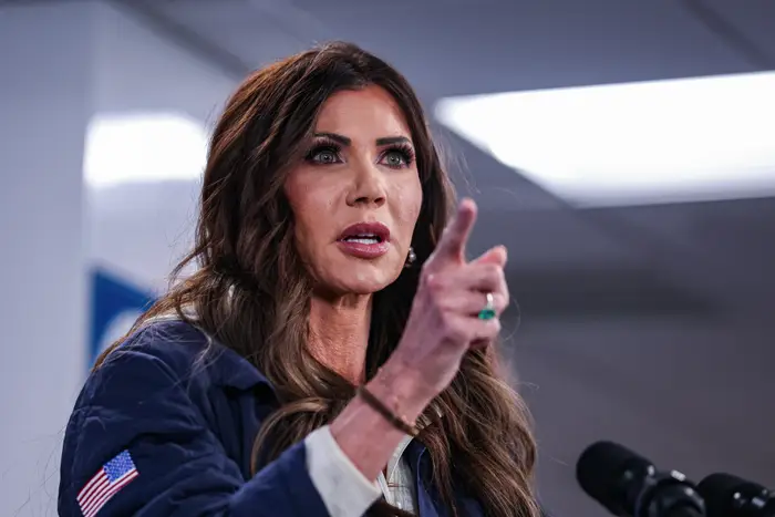 Person som snakker lidenskapelig ved et podium, iført en jakke med flagglapp. Håret er langt og bølget. Mikrofon synlig i forgrunnen