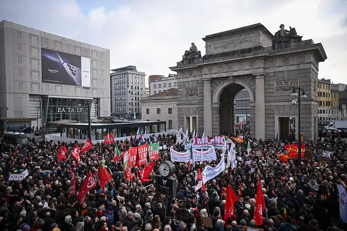Stor folkemengde som protesterer nær en historisk buegang, holder bannere og flagg, med bybygninger i bakgrunnen