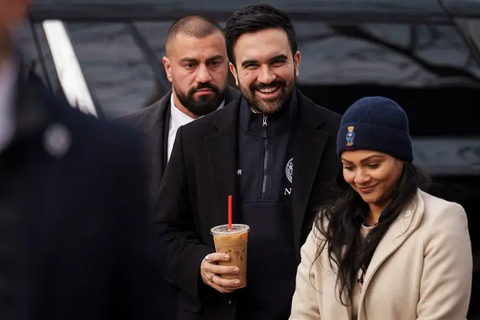 En smilende mann holder en iskaffe ved siden av en kvinne i vinterfrakk og lue. En mann i bakgrunnen har på seg en dress