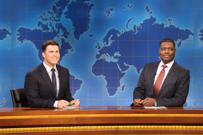 Colin Jost og Michael Che som nyhetsankere ved en pult i SNL-studioet med verdenskart i bakgrunnen, iført dress og slips