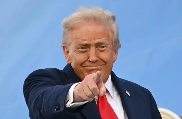 En person i dress peker mot kameraet, smilende lett, foran et nøytralt bakteppe