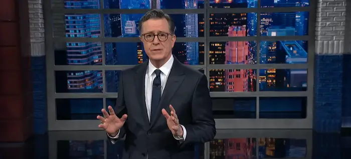 En person i dress gestikulerer mens han er vert for et talkshow, med et bybilde synlig gjennom store vinduer bak seg