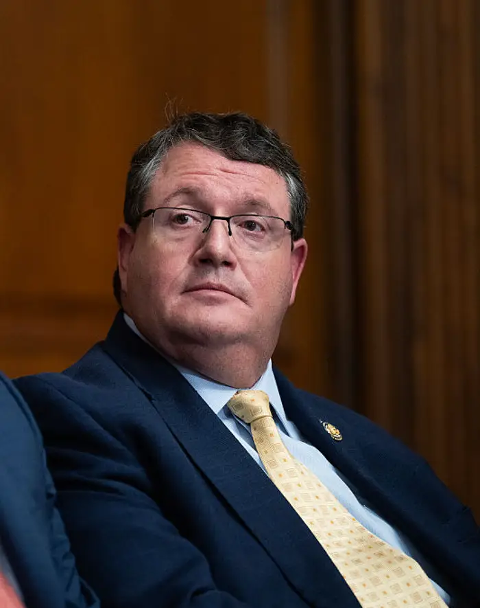 En person i dress og slips sitter oppmerksomt og virker gjennomtenkt under et innendørs arrangement eller møte