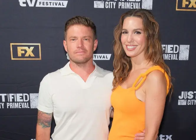 To voksne på en rød løper. Den ene har på seg krempolo og blå bukser; den andre i en gul figursydd kjole. De poserer foran hendelseslogoer