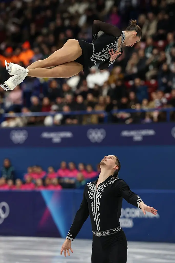 Kunstløpsduo som utfører et løft under en konkurranse, med den kvinnelige skateren i en positur midt i luften, og begge har på seg intrikate mønstrede antrekk