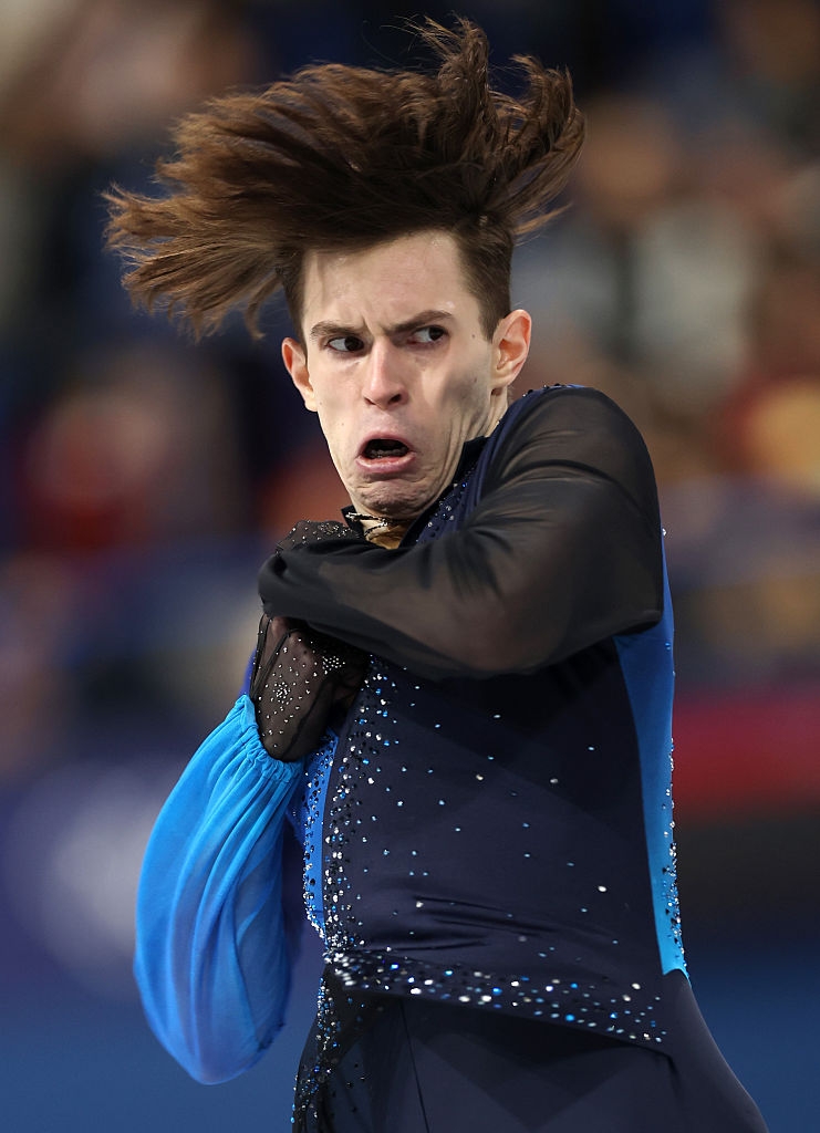 Kunstløper midt i ytelse, intenst ansiktsuttrykk, iført et pyntet antrekk med flytende ermer. Hår fanget i dramatisk bevegelse