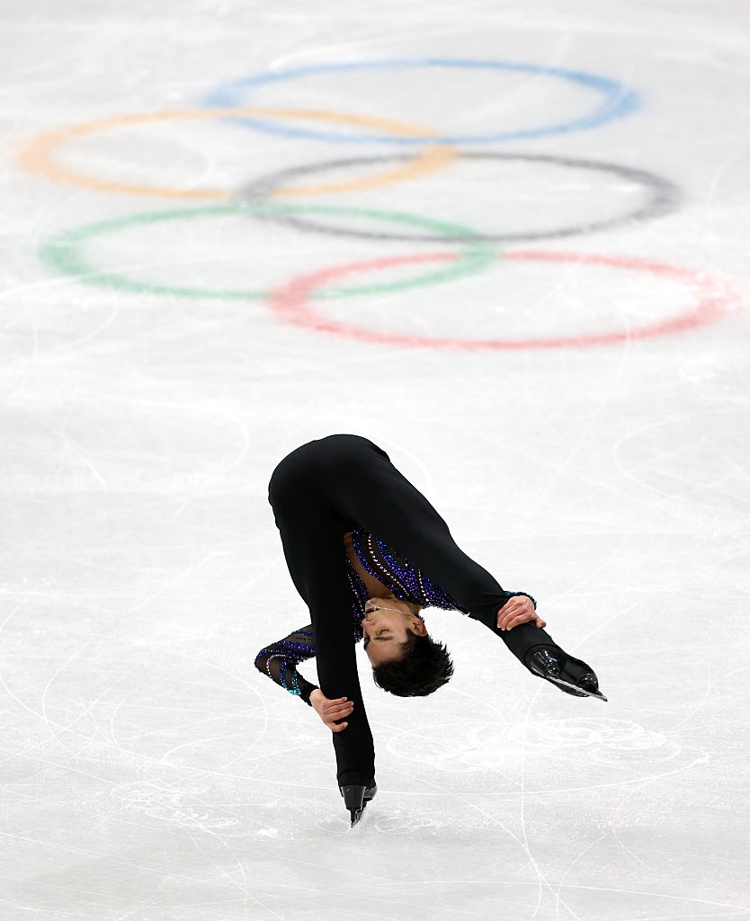 Kunstløper utfører et bakoverbøyende spinn på is, olympiske ringer synlige i bakgrunnen
