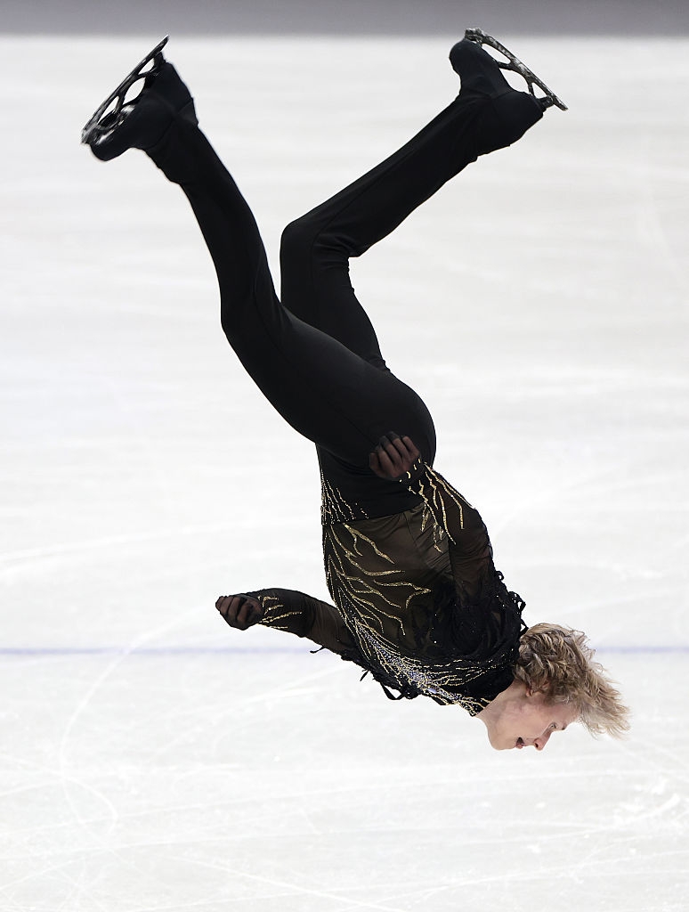 Kunstløper i luften under et hopp, iført et elegant kostyme med glitrende detaljer, på en skøytebane