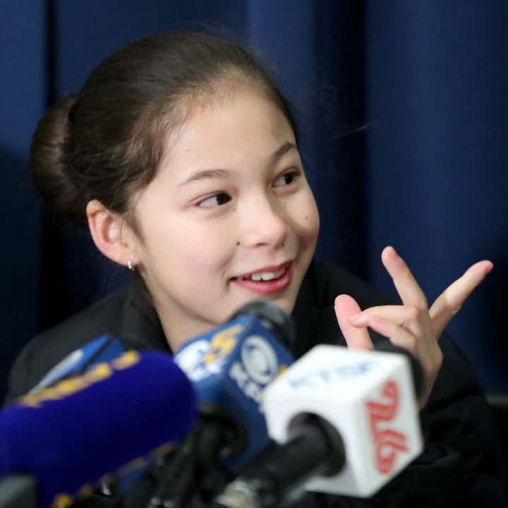 En person som snakker på en pressebegivenhet, smiler og gestikulerer med hånden nær mikrofoner