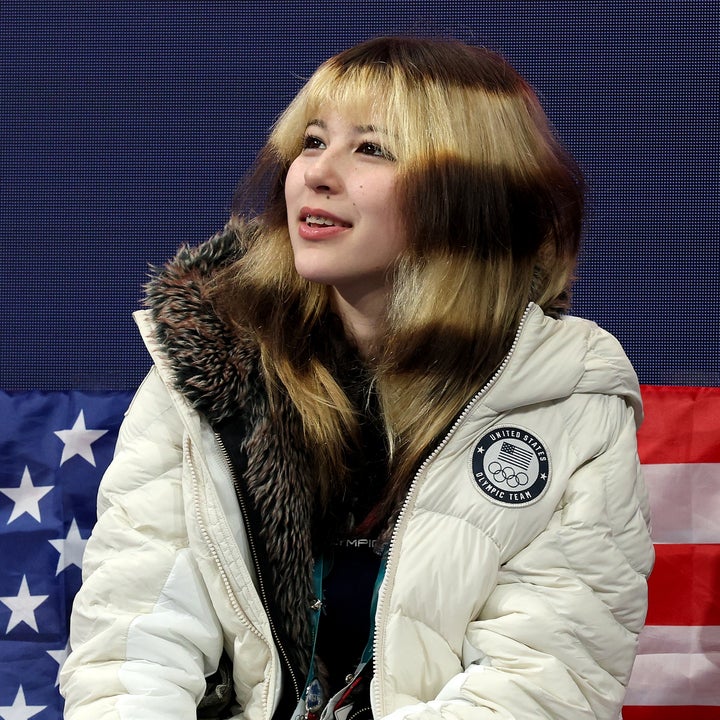 Person i vinterfrakk sitter foran et USA-flagg som bakteppe og ser oppover, muligens på et arrangement eller en konkurranse