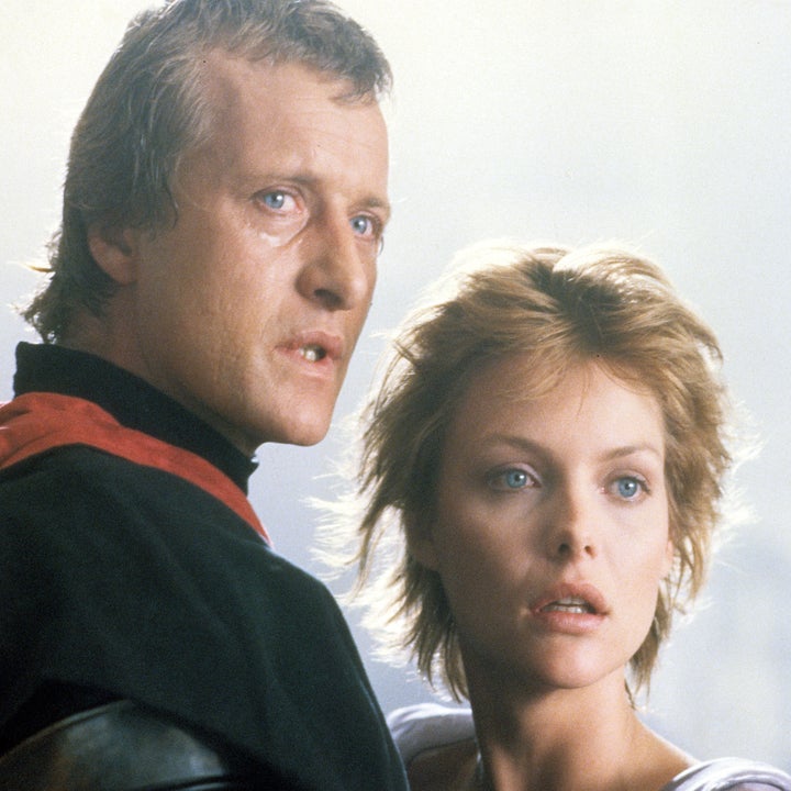 To skuespillere i en dramatisk scene, med alvorlige uttrykk. Mannen har på seg et mørkt antrekk med rød krage; kvinnen har kort hår og et våkent blikk