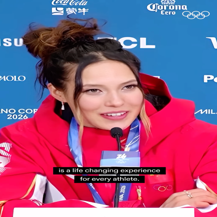 En person i en rød hettejakke snakker på en pressekonferanse med 'Milano Cortina 2026' synlig i bakgrunnen. Bildeteksten lyder: "er en livsendrende opplevelse for hver idrettsutøver."