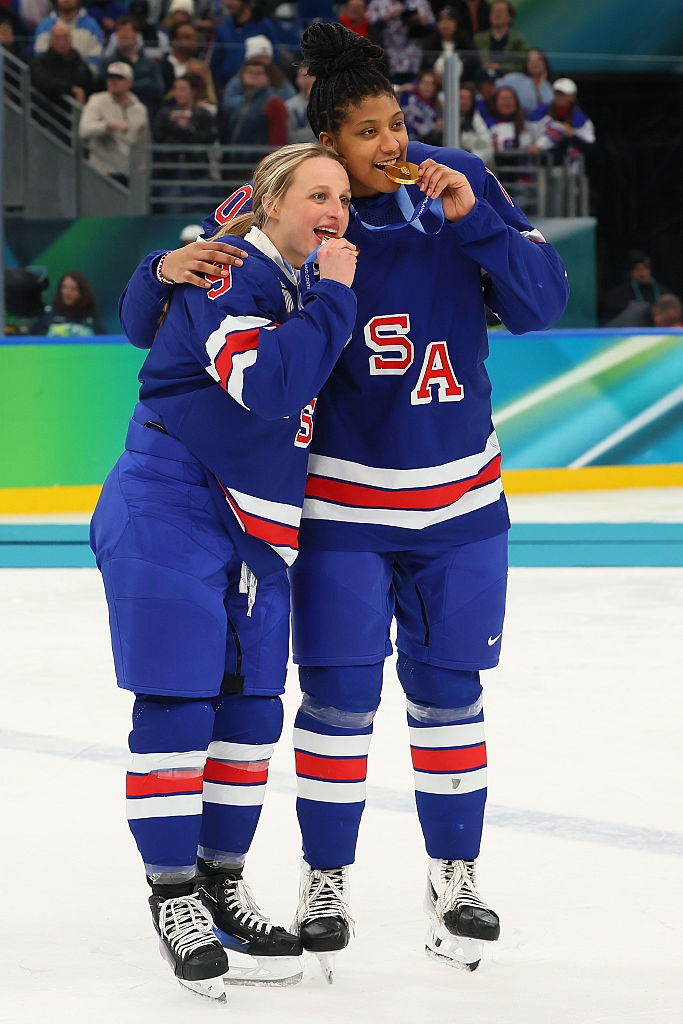 To ishockeyspillere i amerikanske uniformer feirer ved å bite gullmedaljer på isen