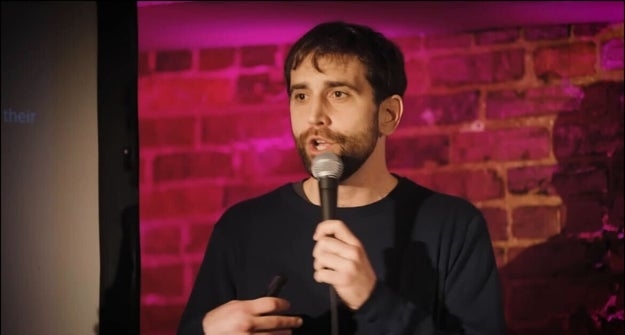 Person som holder en mikrofon, midt i tale, foran en murvegg med lilla belysning. Uformelt antrekk