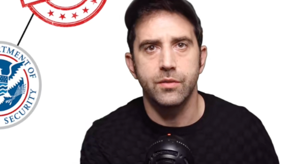 En person i svart skjorte og caps sitter foran en mikrofon med en Department of Homeland Security-logo i bakgrunnen