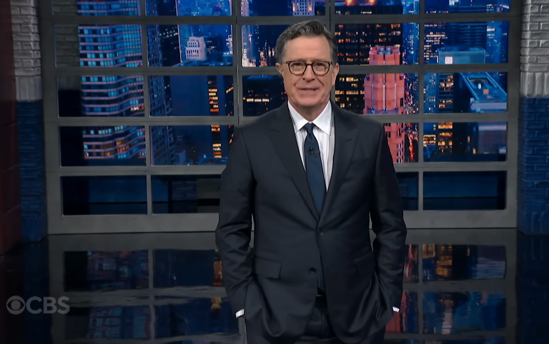 Mann i dress som står på et showsett sent på kvelden, med et bybilde i bakgrunnen