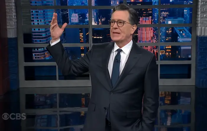 En person i dress holder en monolog på et talkshowsett med et bybilde som bakteppe