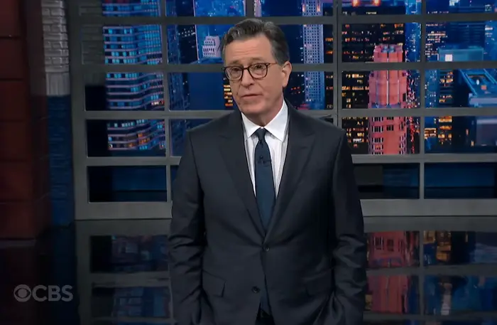 TV-vert står i et studio med bybilde i bakteppe, iført klassisk mørk dress og slips, og fremstår klar til å henvende seg til publikum