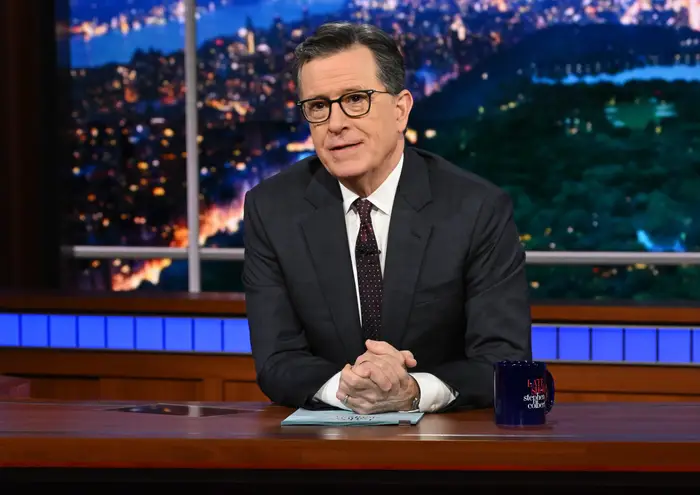 Talkshowvert sitter ved et skrivebord på et sett sent på kvelden, iført mørk dress og mønstret slips, med byens skyline i bakgrunnen